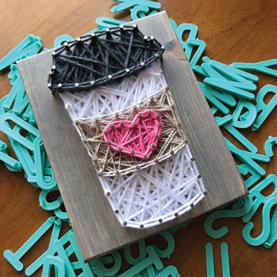 rustic wood plaque with nails and string patterned to resemble a white cup of coffee with black lid pink heart and tan sleeve