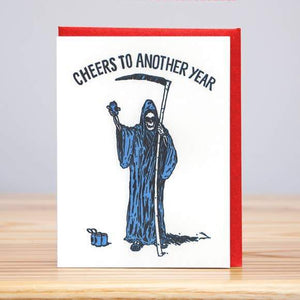 A white card with a text: "CHEERS TO ANOTHER YEAR" and an illustration of a reaper holding a beverage and a scythe. Comes with a red envelope