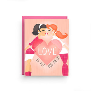 A peach colored card with text: "LOVE IS ALL YOUR NEED" and an illustration of two women kissing. Comes with a pink envelope.