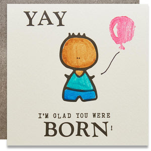 A white square card with a black text: "YAY I'M GLAD YOU WERE BORN!" and an illustration of a boy holding a pink balloon. 